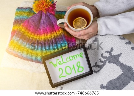 Welcome 2018, text in frame. Winter clothing background and cup of hot tea with lemon.