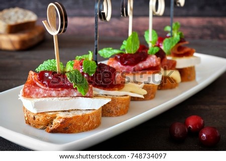Holiday crostini appetizers with cranberry sauce, brie, salami, and mint on white serving plate against rustic wood