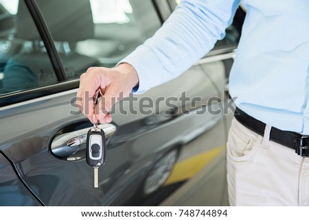 Close up photo of male car dealer hand holding a car key next to a car door. Person is wearing smart casual clothes in beige and blue colors.