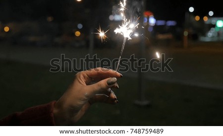 Beautiful sparkler in woman hand on black or street background. Woman holding sparkler against colorful defocused lights, close up. Female hands holding sparkler on dark background