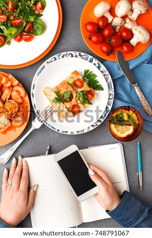 Unrecognizable woman have a lunch break eating casserole and using smartphone on gray background, copy space on screen. Healthy eating concept, organic vegetables top view.