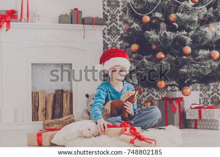 Cute happy boy in santa hat holding toy. Christmas present on holiday morning in beautiful room. Male child got Xmas gift near decorated fir tree and fireplace. Winter holidays concept