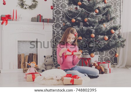 Cute happy girl unwrap christmas present box on holiday morning in beautiful room interior. Feale child open Xmas gifts near big decorated fir tree and fireplace. Winter holidays concept