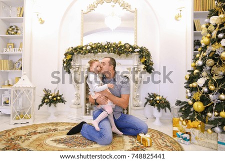 Adult man and young father playing with girl, tickles and kisses his daughter, laughing girl kneeling on Pope against fireplace with candles decorated Christmas tree and yellow balls and mirrors. Men