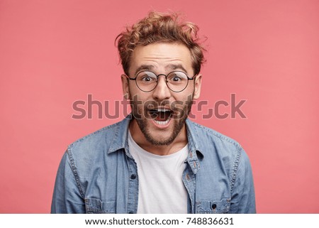 Excited bearded man wears glasses and denim shirt watches football match with friends, being great fan, involved in game, screams happily because of scored goal, rejoice team`s victory and success