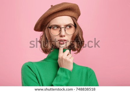Indoor shot of pensive young female teacher in green polo neck sweater, beret and round spectacles looks thoughtfully aside as plans interesting lesson for talented pupils, generates creative ideas.