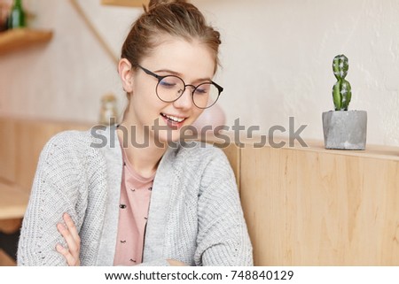 Delightful dreamy woman in eyewear sits near wooden wall of home or cafe interior, looks down, reads newspaper or magazine with attentive look, enjoys free time and calm domestic atmosphere.