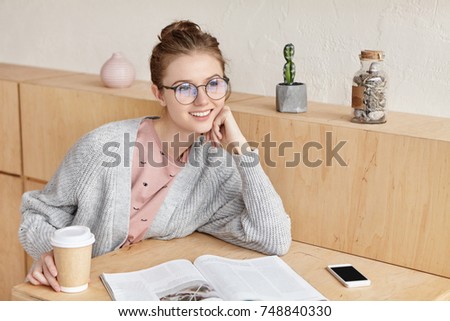 Female student wears loose sweater and round glasses, reads book as waits for groupmate in cafeteria, drinks takeaway coffee, has dreamy expression, sits at wooden table, uses modern gadget.