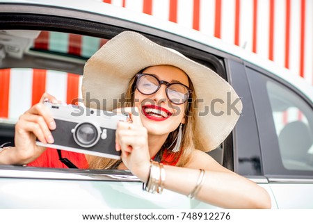 Poortrait of a young happy woman pulling out of the car window with photo camera on the red wall background during the summer vacation