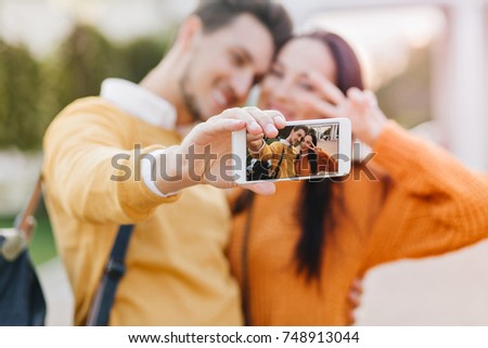 Cute girl posing with peace sign while her boyfriend in orange sweater making selfie. Funny couple using smartphone for outdoor  photoshoot in autumn day.