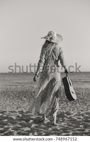 Attractive woman holding guitar on sunset sea beach outdoors background. Back side view of emotional walking female, instrumental relaxation