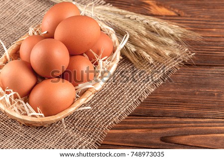 Red chicken eggs in straw basket and wheat ears on piece of sacking on brown wooden textured background - eco farming concept. Close up photography of organic food.