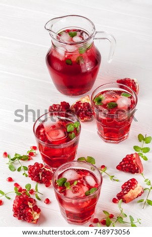 Pomegranate cocktail with ice and mint in beautiful glasses and jug with fresh ripe fruit on white wooden background. Close up photography of sweet red juice.