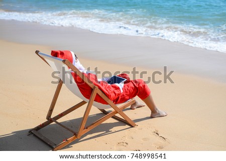 Santa Claus using tablet pc on beach tropical background. Modern internet technology holidays. Back side view of person doing xmas education project, take self photo, relaxing, play gadget or shopping
