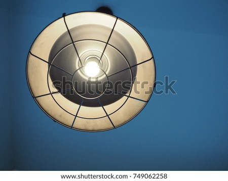 A round big industrial black and white lamp hanging from the blue ceiling with single lighting bulb and its warm light shining around