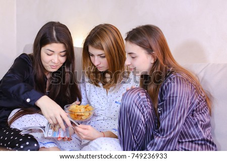 Three wonderful women together hold fashionable women's magazine in their hands and read articles or view bright pictures, eat potato chips, chat and gossip, sitting on gray sofa in bright bedroom at