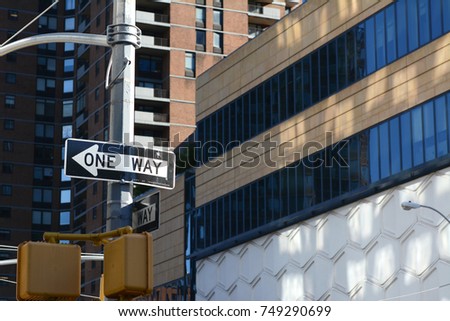 ONE WAY road sign points left on a New York City street, with high rise buildings beyond