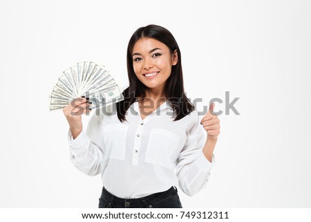 Picture of smiling young asian woman standing isolated over white background. Looking camera holding money showing thumbs up.
