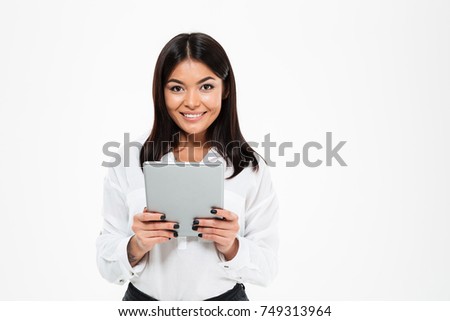 Picture of happy young asian lady standing isolated over white background chatting by tablet computer. Looking camera.