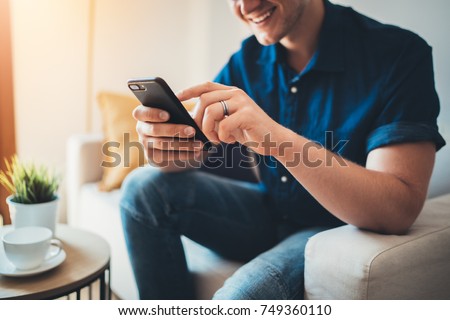 Happy smiling young man using modern smartphone device while sitting on sofa at home, modern design interior, cheerful hipster guy typing an sms message at social network Royalty-Free Stock Photo #749360110