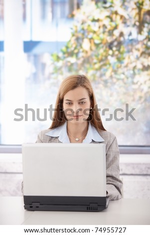 Attractive young businesswoman sitting at desk, working on laptop in bright office.?