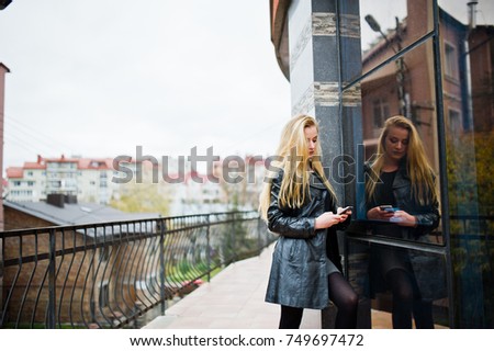 Blonde fashionable girl in long black leather coat posed against large window of building.