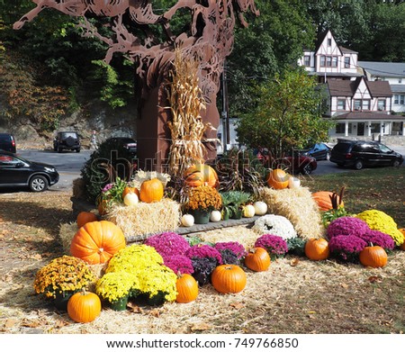 bright colored mum flowers, orange pumpkins and hay bales by a tree.  The ground has fallen leaves.