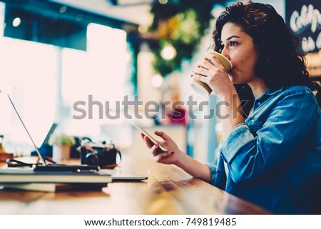Attractive smart graphic designer drinking aroma coffee while distantly working on gadget in cafe.Young female person enjoying beverage while transferring money online via banking service on telephone