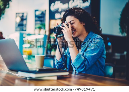 Skilled female photographer taking picture on cool vintage camera while sitting at wooden table indoors.Young woman trying to make good photo using retro equipment during leisure time in cafe