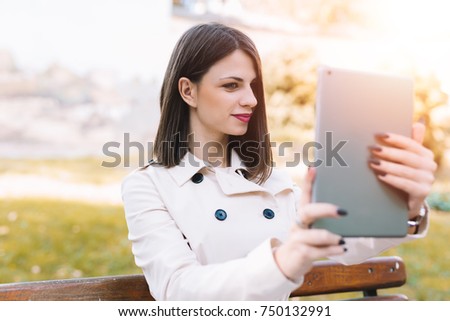 Attractive young girl taking selfie in the park with her tablet. Sitting on the bench
