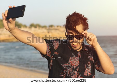 The ugly man wears blue flowery shirt and glasses on sunny and hot day at the beach. Funny man doing selfie with his smart cell phone.