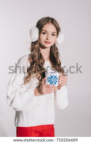 beautiful teenage girl in fur earmuffs holding cup and looking at camera isolated on white