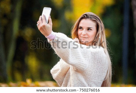 Woman in warm sweater taking picture by mobile phone walking in the autumn park