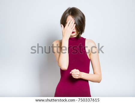 beautiful young woman hides face with hands, dressed in a burgundy dress, isolated in studio on a gray background