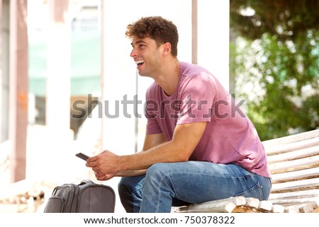 Side portrait of happy man sitting on park bench with suitcase and cellphone