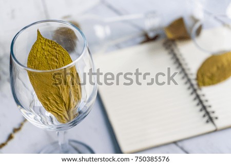 A beautifully decorated wine glass with brown leaves around it and a diary placed on a white background makes it beautiful and attractive.
