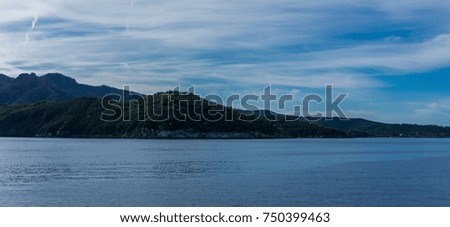 Horizontal photo on cliff which goes deep to the Mediterranean sea. The rocks and mountains of Elba island are covered by green bush and trees. Sky is blue with clouds.