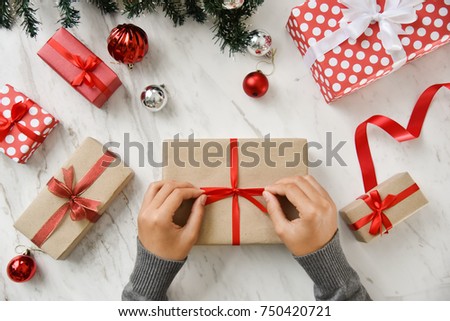 Female hands wrapping a brown present box with red ribbon on white marble background for Christmas and New year. Top view.
