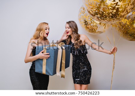 Gorgeous woman with elegant hairstyle holding big gift with surprised face expression. Indoor photo of two pretty girls having fun during celebration and posing on light background.