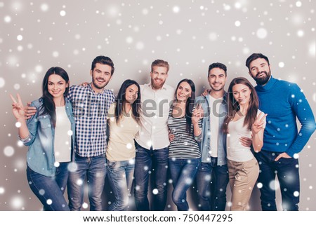 In winter eight cheerful young people embracing and smiling in casual clothes. Ladies are showing two fingers sign, snowflakes background