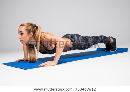 Young attractive woman practicing yoga, doing Push ups or press ups exercise, Plank pose, working out, wearing sportswear, red tank top, pants, indoor full length, isolated, grey