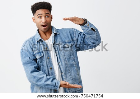 Isolated portrait of young funny dark-skinned man with afro hairstyle in casual white shirt under denim jacket pretending holding big box in hands with excited face expression