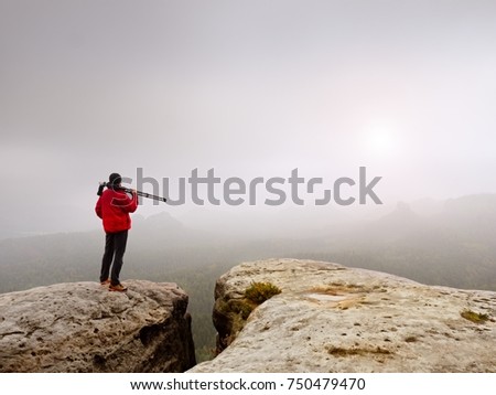 Hiker with camera on tripod takes picture from rocky summit. Alone photographer at edge photograph misty landscape, forest in valley. 