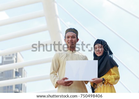 Young Arab man and young Arab woman holding a white blank horizontal board at front modern building in city.