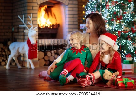 Family with kids at Christmas tree and fireplace. Mother and children opening gifts at fire place. Boy, girl and mom open presents. Winter holidays interior decoration. Child in pajamas on Xmas eve.