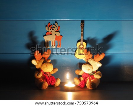 Two toy Christmas moose, or elks, are sitting on a background of blue wall with a candle, a guitar and romantic silhouettes in wooden rural house; medium shadows