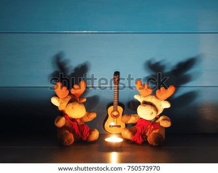 Two toy Christmas moose, or elks, are sitting on a background of blue wall with a candle, a guitar in wooden rural house; medium shadows