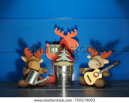 Two toy Christmas moose, or elks, sitting by a small Christmas tree near the blue wall with a candle, a guitar and romantic silhouettes in wooden rural house; medium shadows