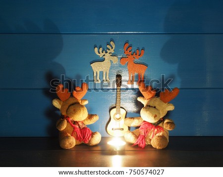 Two toy Christmas moose, or elks, are sitting on a background of blue wall with a candle, a guitar and romantic silhouettes in wooden rural house; very big shadows