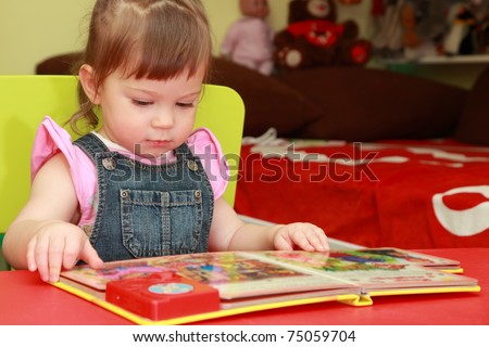 beautiful little girl in denim jumpsuit and pink shirt seats on yellow chair and reads book